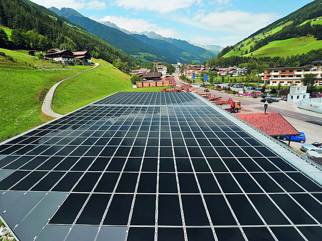 A solar plant on the roof of a building produces electricity, with green alpine slopes rising to the left and right.